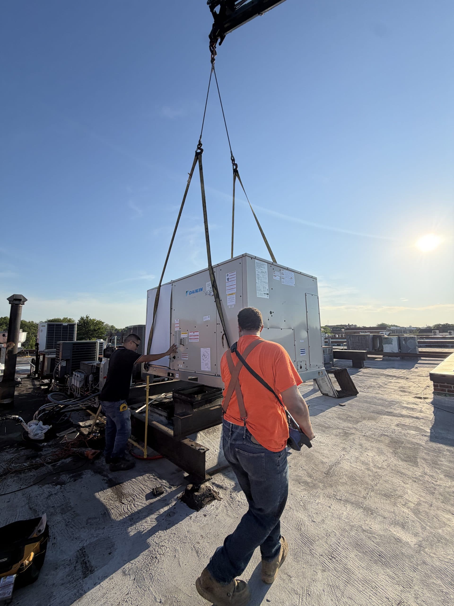 Vinco crew rigging a Daikin R-32 rooftop unit with a crane, rigging straps visible against the sky